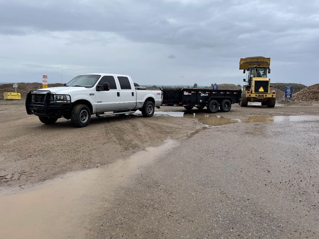 White Ford F-250 with black dump trailer and a yellow loader at a materials yard